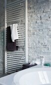 Towels hung on a white hand towel heater on the wall with decorative facing strips with a natural stone look with a partially visible bath tub in the foreground