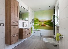 Modernes Bad mit erfrischendem Naturmotiv auf grossformatiger Glasplatte an Wand im Duschbereich, an der Seite montierter Hochschrank mit Holzdekor neben Waschtisch mit Schubladenschrank und gleichem Holzdekor
