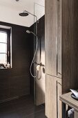 Bodenebene Dusche mit feststehender Glas Abtrennung, Kopf- und Handbrause an gefliester Wand, im Vordergrund Hochschrank mit Holzdekor Eiche und Edelstahl Griffleisten