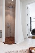 An oval rain shower cubicle with a limestone mosaic wall, a teak floor grating and a curtain in a cosy en suite bathroom with a view through a frame and panel door into the bedroom