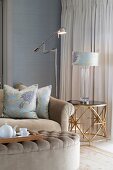 Teepause in elegantem Wohnzimmer - Tablett mit Teegeschirr auf Polstertisch, Sofa mit eleganten Kissen, passende Tischleuchte auf Beistelltisch und Stehleuchte aus Edelstahl