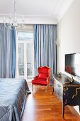 Antique-style chest of drawers, Rococo-style, red armchair and pale blue curtains at balcony windows in traditional bedroom