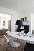 Black pendant lamp above white, oval table an shell chairs; piano and sofa in adjoining living area in background