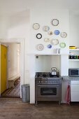 Various ornamental wall plates above stainless steel gas cooker in disused fireplace niche of period-apartment kitchen; view into hallway with yellow door