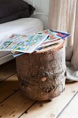 Comics on tree stump table on castors used as bedside table, stripped wooden floor, metal bed and floor-length linen curtains