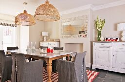 Wooden table with marble top and chairs with grey loose covers below wicker pendant lamps in rustic dining room