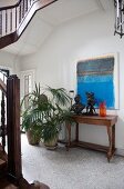 Stairwell with pale grey, mosaic floor tiles, antique console table and potted palms below modern artwork on wall