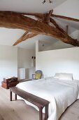 Double bed against partition and simple wooden bench at foot of bed in white bedroom with rustic, wooden ceiling beams