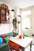 Gemütliche, kleine Wohnküche mit farbigen Stühlen an antikem Holztisch, dahinter ein Vintage Schränkchen über Truhenbank mit Kissen