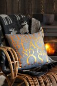 Scatter cushion with gold lettering on grey background and reindeer-patterned blanket on wicker armchair in front of wood-burning stove