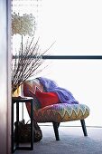 Rotes Kissen auf Sessel mit bunt gestreiftem Zickzackmuster, daneben Zweige in Vase auf Beistelltisch und Designerlampe