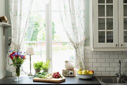Vegetables on chopping board and vase of flowers on kitchen worksurface in front of window with gathered curtains