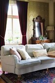 Pale couch with scatter cushions in front of red, gathered curtains on windows in traditional living room