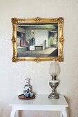 Flask and beakers on tray next to vintage oil lamp on white console table below gilt-framed picture on wall