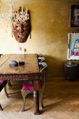 Hand-crafted Oriental wooden mask on gold wall, chandelier and carved exotic-wood table in eclectic interior