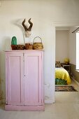 Various containers on top of pink-painted wooden cupboard below hunting trophy on wall next to open bedroom door