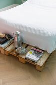 Mit Waffelpiquee-Decke bezogenes Bett auf überstehender Europalette als Nachttisch