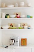 Weisses und pastellfarbenes nostalgisches Geschirr auf Regalböden an weisser Küchenwand mit Küchenmaschine auf Marmor-Küchenarbeitsplatte