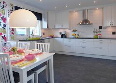 White chairs at set table in front of fitted kitchen with white cabinet fronts