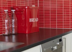 Empty milk bottles and red metal bread bin on kitchen worksurface with splashback of red mosaic tiles