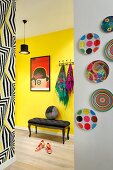 Framed red poster above antique, upholstered bench against yellow hallway wall, colourful plates on white wall and wall with patterned wallpaper