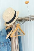 Straw hat and denim jacket on small, stainless steel coat rack on pale wooden wall