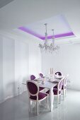Elegantly set dining table and crystal chandelier in dining room with purple, indirect lighting on ceiling and Neo-Baroque, upholstered chairs