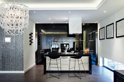Black bar stools at counter in open-plan kitchen in modern interior with indirect ceiling lighting and chandelier to one side
