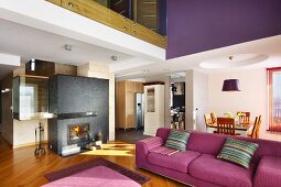 Modern violet couch in open-plan interior with dining area and fireplace in partition wall below mezzanine