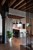 Industrial charm in loft apartment with black metal column under concrete girder and wood-beamed ceiling; modern dining area with row of pendant lamps in background