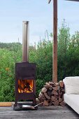 Fire in log-burner next to small pile of firewood on terrace, pale couch to one side and view into densely planted garden