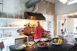 Woman standing at cooker in open-plan kitchen; bowls of fruit on black stone surface of island counter in foreground