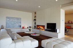 Interior with white designer sofa and set of coffee tables in front of fireplace in wall with shelf niches and masonry shelf running around two walls