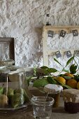 Pears under glass cover, freshly picked lemons and jars of jam in front of old wooden frames on roughly plastered wall