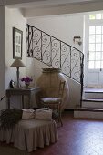 Small antique desk and chair, huge old olive amphora and bunch of lavender on pouffe on terracotta tiles at foot of staircase with wrought iron balustrade