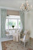 Chandelier above elegant seating area with delicate, white antique furniture in front of traditional, Scandinavian window with curtains