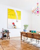Cane cantilever chairs around dining table below colourful chandelier with multicoloured glass droplets in front of modern painting on wall