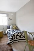 Black and white bedspread with geometric pattern on bed next to retro standard lamp in front of window; green metal chair in foreground