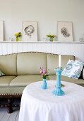 Cottage Flair, runder Tisch und antikes Sofa vor weiß lackierter Holzverkleidung mit aufgestellten gerahmten Zeichnungen