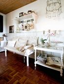 Serving trolley next to white bench with scatter cushions and bolsters against wainscoting in rustic interior with mosaic parquet floor