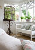 Vintage bird cage with ornate metal stand next to white bench in wood-clad conservatory