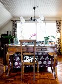 Orchids on antique mahogany desk and chair in rustic living room with white clad, sloping ceiling