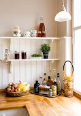 Fruit bowl next to tray of condiments on kitchen counter below bracket shelves on white wooden wall