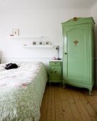 Rustic corner cupboard painted green, bedside cabinet and cat on double bed with patterned bedspread