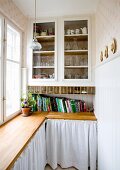 Fitted cabinet with glass doors above wooden worksurface with white curtains below in pantry