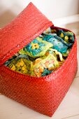 Red-painted rattan basket with open lid showing colourful crocheted squares with floral patterns