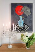 Modern artwork on wall above lit candles in crystal candelabra and vase of hydrangeas on sideboard
