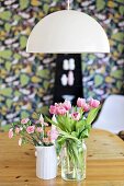 Pink tulips and carnations in vases on wooden table below pendant lamp with white, hemispherical lampshade
