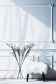 Wall with decorative strips, in front of it stool with white fur cover and branches in a glass vase