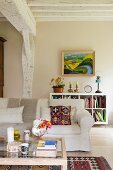 Coffee table, sofa with white loose cover and painting on wall above low shelving unit in interior with whitewashed wooden beams